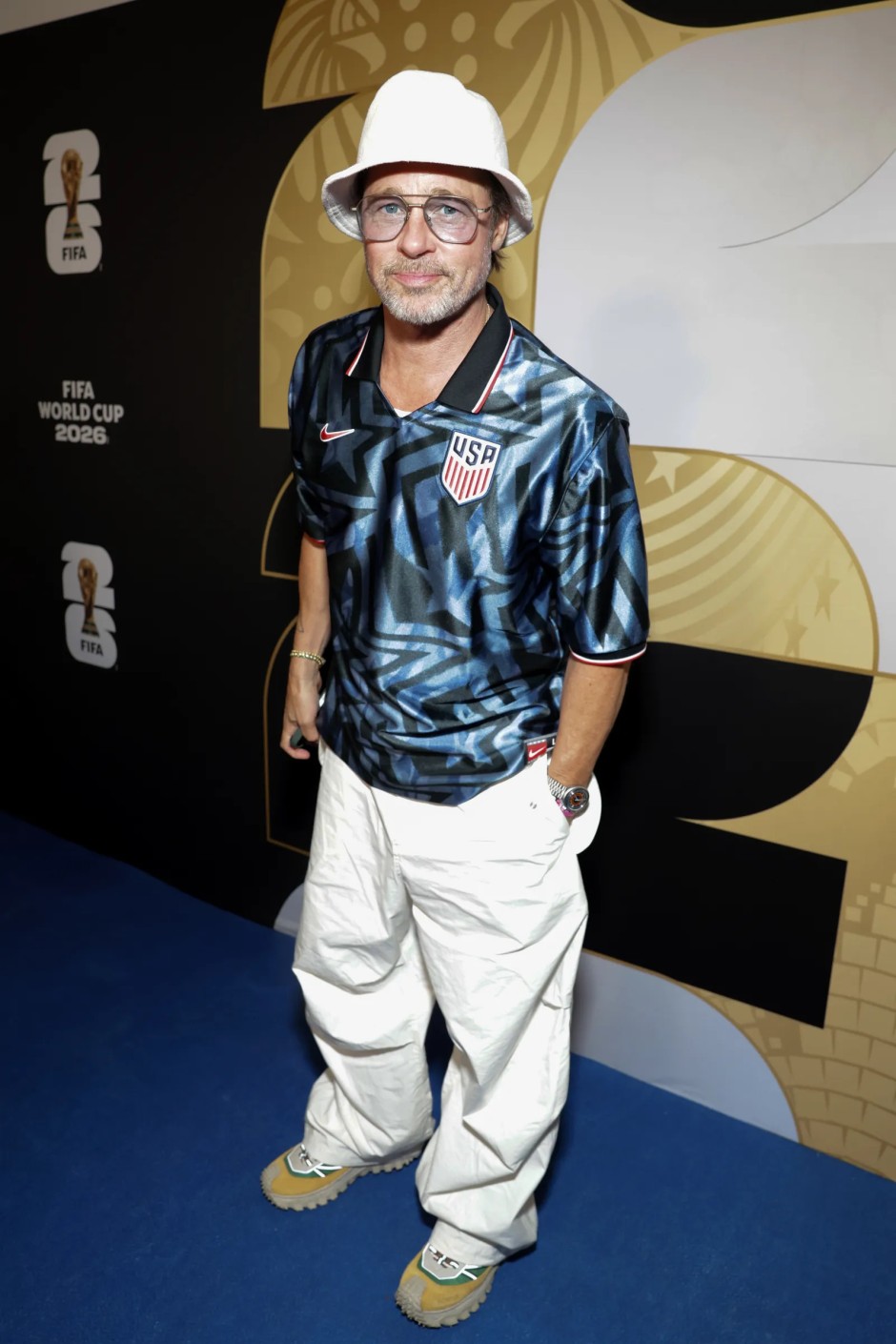 Brad Pitt, ABD Milli Takımı formasını beyaz bucket şapka, bol beyaz pantolon ve spor ayakkabılarla kombinleyerek FIFA Dünya Kupası 2026 etkinliğinde poz veriyor.