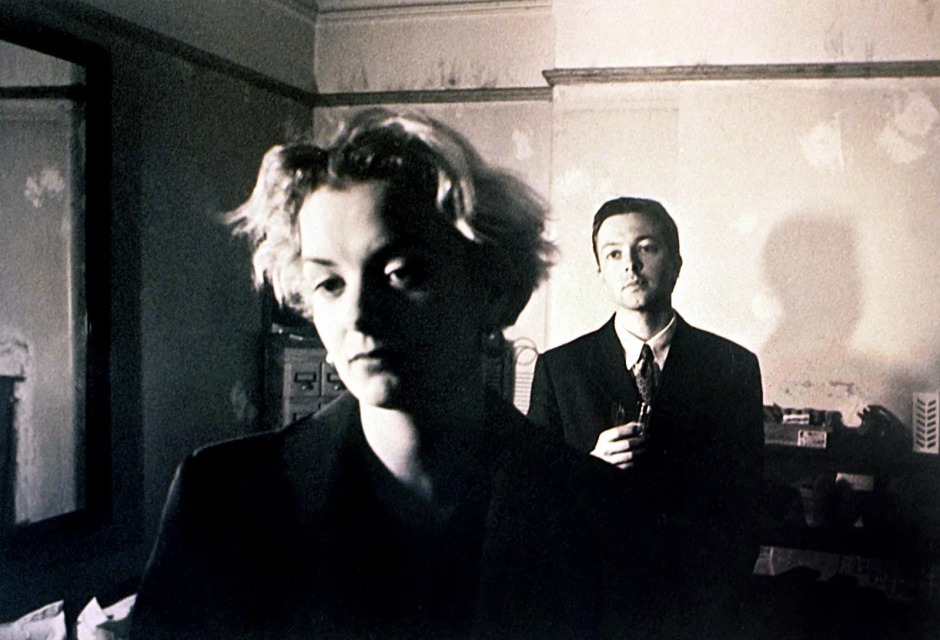 Christopher Nolan'ın Following (1998) filminden siyah-beyaz bir kare; ön planda genç bir kadın, arka planda takım elbiseli bir erkek karakter yer alıyor.
